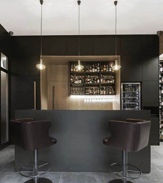 Modern man cave with two black bar stools, a minimalist black counter, and shelves stocked with bottles and glassware