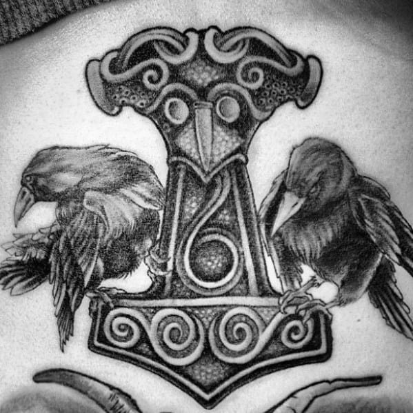 Black and grey tattoo of Huginn and Muninn perched on Mjölnir, Thor’s hammer, with intricate Norse detailing and shading.