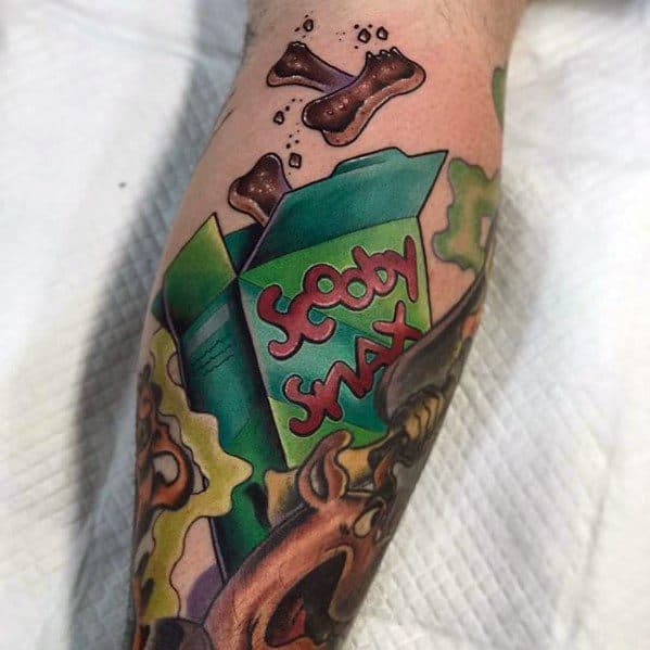 Tattoo of a green Scooby Snacks box with bones flying out, surrounded by Scooby-Doo and colorful swirling background elements.