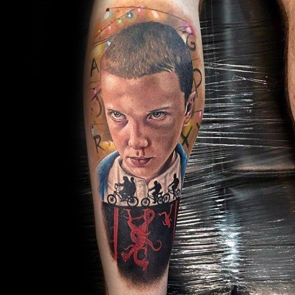 Realistic tattoo of Eleven from Stranger Things with the alphabet wall above and the kids biking above the Upside Down below.