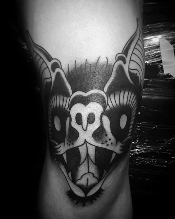 Black and grey bat head tattoo with glowing eyes, bold shading, and exaggerated features placed on the back of the knee.