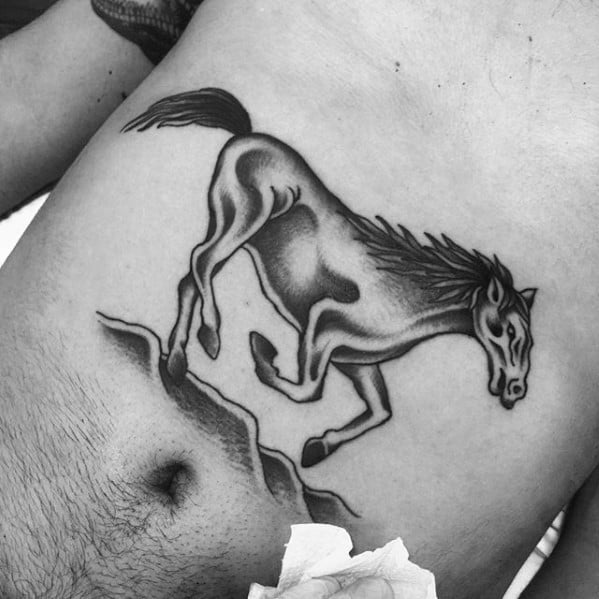 Black and grey stomach tattoo of a galloping horse with flowing mane, placed above the hip and shaded for a dynamic effect.