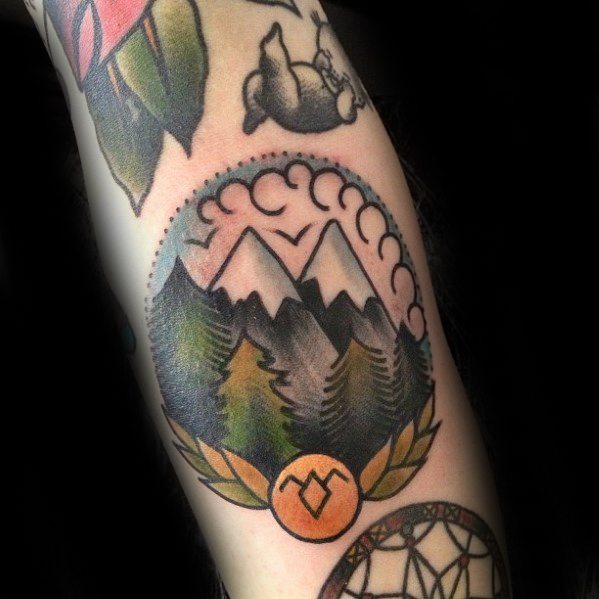 Mountain landscape tattoo with layered pine trees, curved clouds, and a sun symbol framed by leaves and a dotted semicircle.