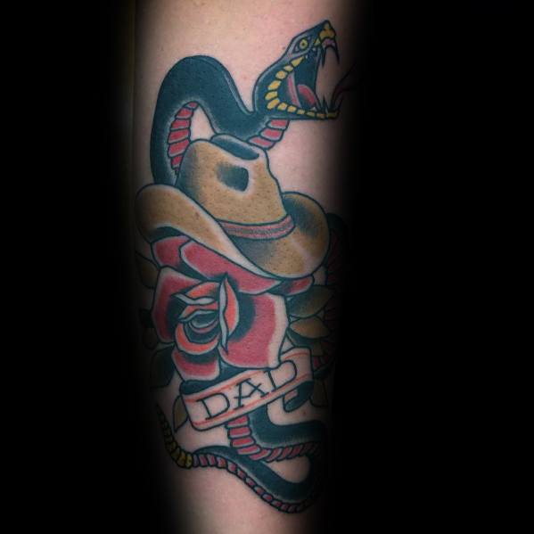 Tattoo of a cowboy hat over red roses with a coiled snake, featuring a "Dad" banner, done in bold traditional style with strong outlines and shading.