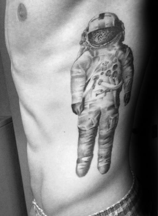 Black and grey Deja Entendu astronaut tattoo on the ribcage with detailed shading and a galaxy inside the helmet.