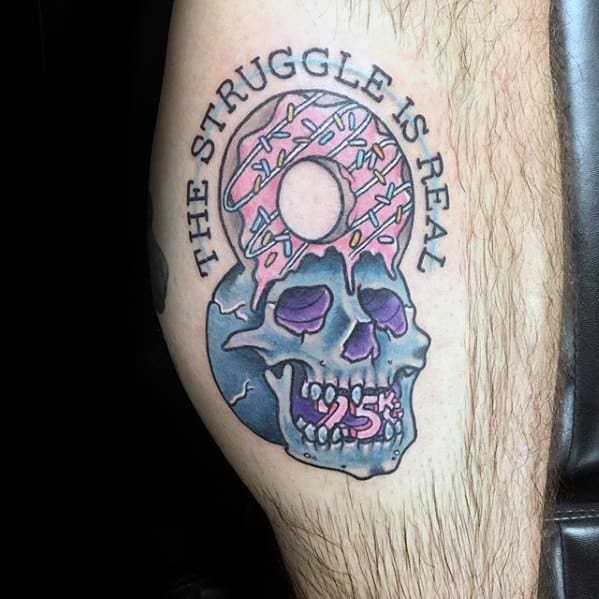 Tattoo of a skull with a pink sprinkled donut on top and the phrase the struggle is real curved above, mixing humor and dark art.
