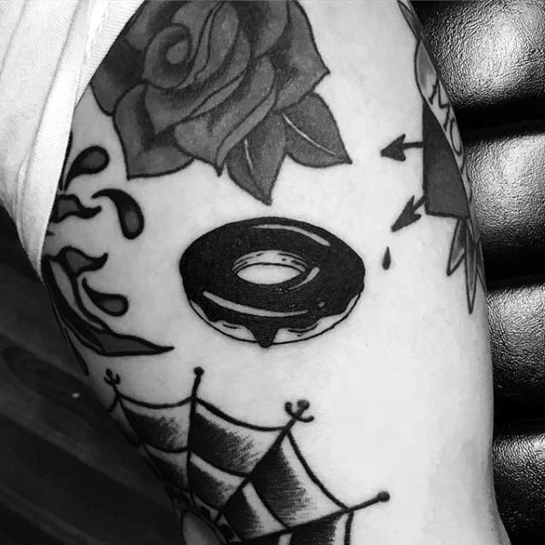 Blackwork tattoo of a simple donut with a bite mark, surrounded by classic bold-style elements like a rose and banner.