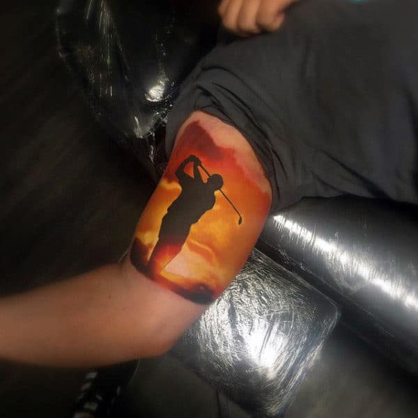 Tattoo of a golfer's silhouette against a sunset on an upper arm