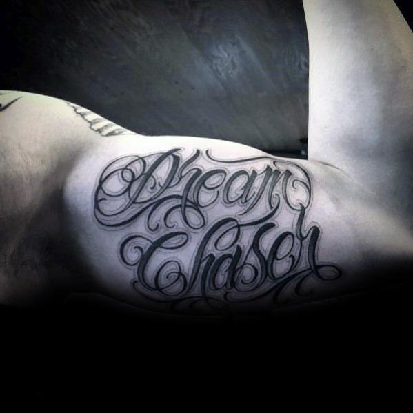 Inner bicep tattoo with the phrase “Dream Chaser” in bold, flowing script, shaded for depth and dramatic contrast.