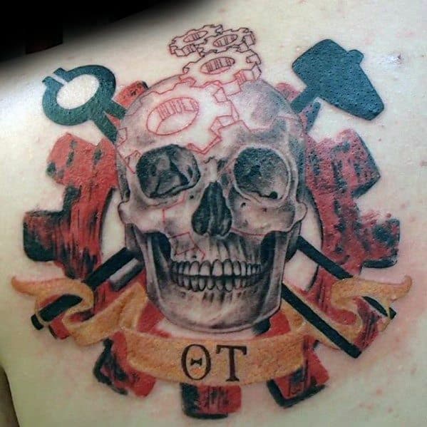 Tattoo of a skull with mechanical gears in the cranium, crossed wrench and hammer behind, set against a red gear and banner marked “OT.”