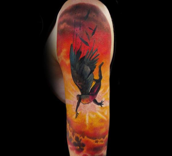 Colorful tattoo of a winged figure falling through a fiery sky, with feathers scattering against a sunset backdrop on the upper arm.