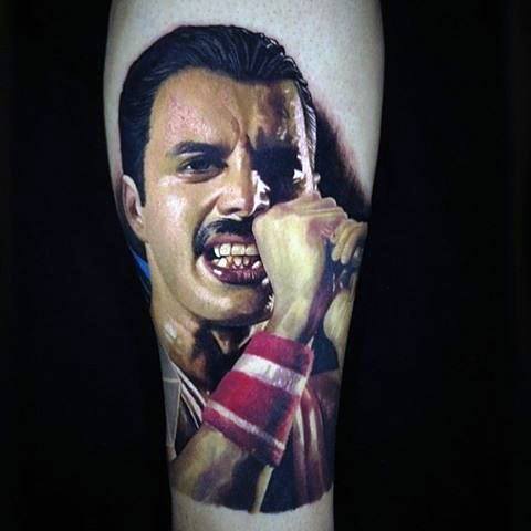 Hyper-realistic color tattoo of Freddie Mercury mid-performance, showing intense emotion and wearing red wristbands.