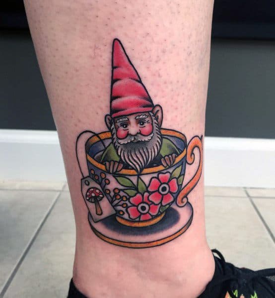 Tattoo of a cheerful gnome with a red hat and white beard, sitting inside a floral teacup with a mushroom tea bag tag, blending whimsy and fantasy.