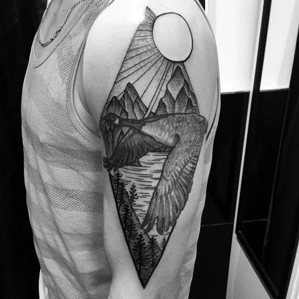 Black and gray tattoo of a flying goose within a geometric diamond frame, featuring mountains, a sun, and a lake, inked on the upper arm.