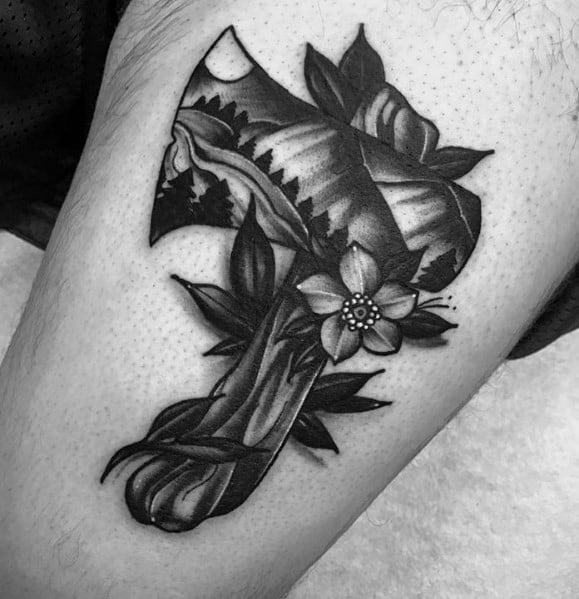 Black and grey hatchet tattoo featuring a detailed mountain landscape on the blade, with floral and leaf accents wrapping around the handle.