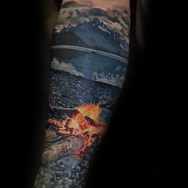 Hyper-realistic campfire tattoo on a rocky lakeshore with detailed flames, wood, and a mountain landscape under a dramatic cloudy sky.