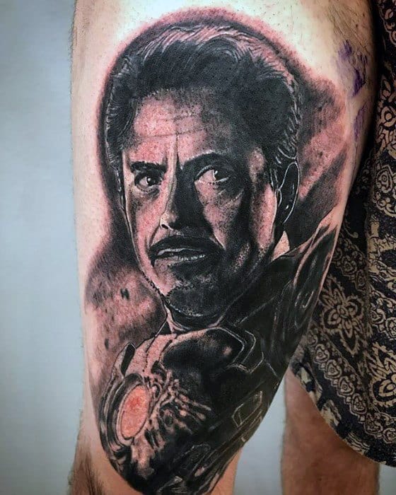 Iron Man tattoo featuring a realistic black and gray portrait of Tony Stark, focusing on his facial expression and the arc reactor, showcasing intense shading and fine detail.