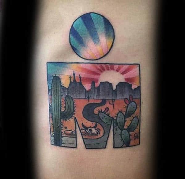 Colorful Ironman logo tattoo featuring a desert landscape with cacti, a winding road, and a sunrise with striped rays in the circle above.