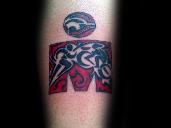 Ironman logo tattoo with tribal-inspired figures of swimming, cycling, and running in black over a red flame-textured background.