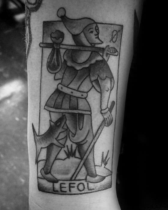 A black and gray tattoo of a jester in a card-like design, walking with a staff and a small dog at his side, with "LEFOL" written below, placed on the forearm.
