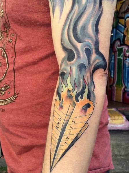 Tattoo of a burning paper airplane on an arm with smoke and flames creating a fiery effect, symbolizing the fleeting nature of adventure and embodying both daring and fragility