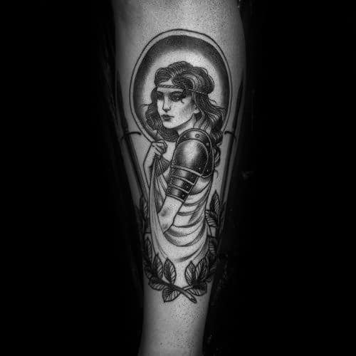 Elegant black and grey Joan of Arc tattoo with soft shading, medieval armor, floral border, and a solemn, introspective expression.