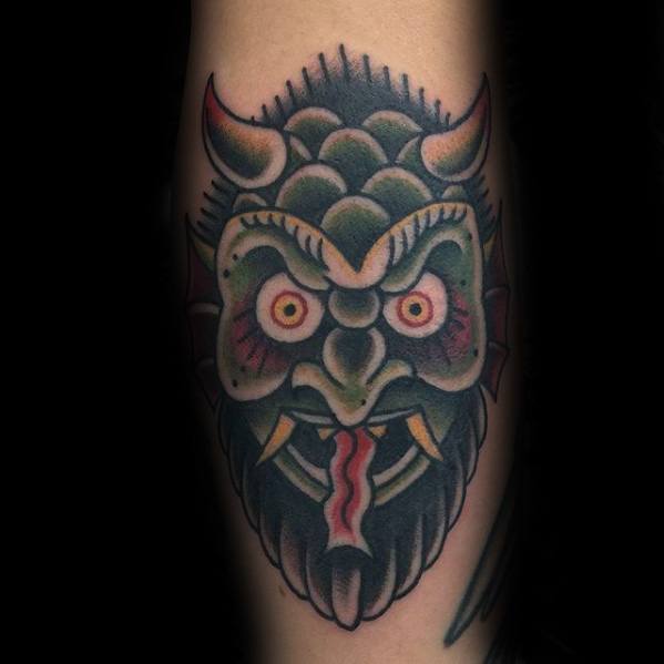 Traditional Krampus tattoo with a horned green and black face, bulging red eyes, sharp yellow fangs, and a red tongue sticking out.