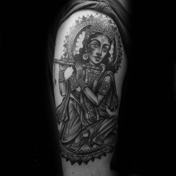 Detailed Krishna tattoo on the upper arm showing him seated and playing the flute, surrounded by an ornate circular halo and traditional attire.