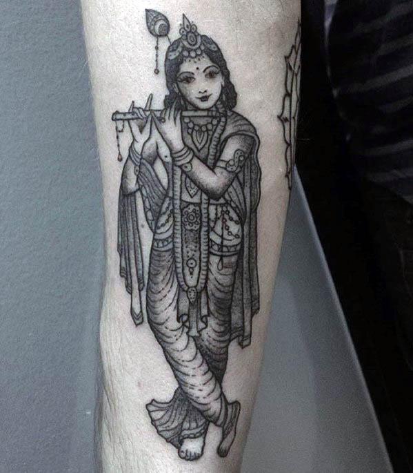 Detailed black and grey Krishna tattoo showing him playing the flute in a classic cross-legged pose with ornate traditional attire.