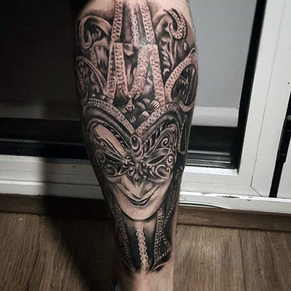 A black and gray tattoo of a detailed jester mask with intricate designs and flowing elements, placed on the calf.