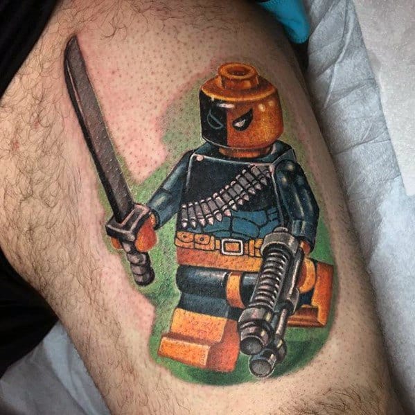 Colorful tattoo of a Lego Deathstroke figure holding a sword and blaster, with vibrant orange, blue, and green tones and detailed shading.