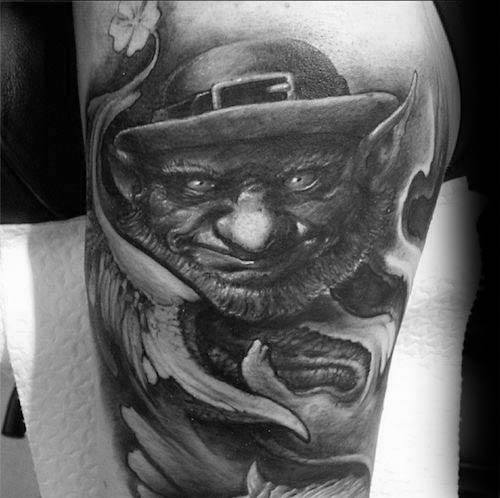 Black and grey tattoo of a mischievous leprechaun with pointed ears, glowing eyes, and a sly grin, surrounded by swirling smoke and a shamrock.