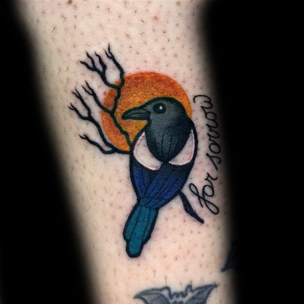 Small magpie tattoo with bold black, white, and blue colors, set against an orange sun with a bare branch and "for sorrow" text on the side.