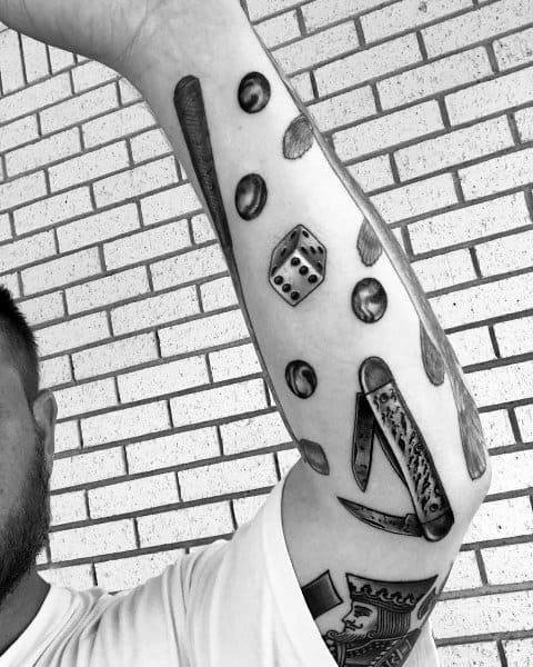 Black and gray forearm tattoo featuring marbles, a dice, a pocket knife, and other vintage objects, creating a nostalgic and detailed design.