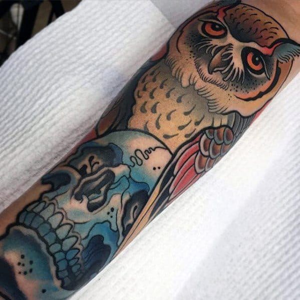 Neo-traditional owl tattoo with piercing orange eyes, bold shading, and a blue skull accent, creating a striking forearm design.