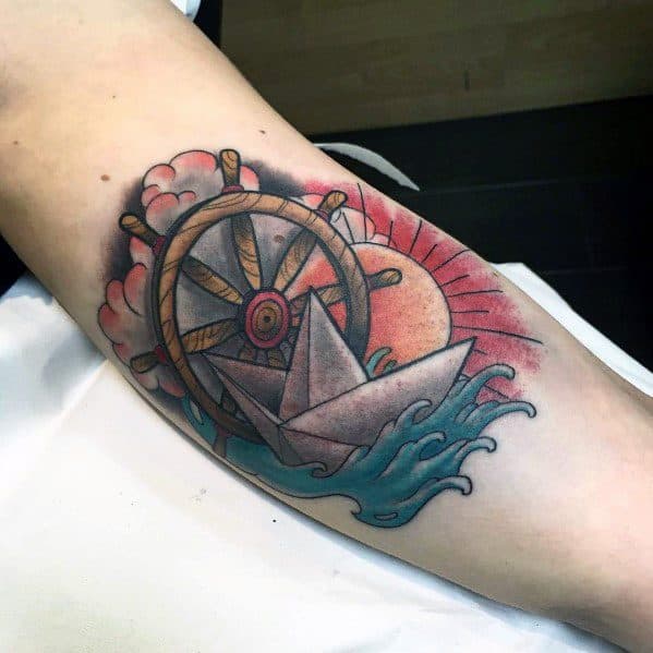 Vibrant paper boat tattoo with a ship’s wheel, waves, and sunset, symbolizing adventure, guidance, and life’s journey.