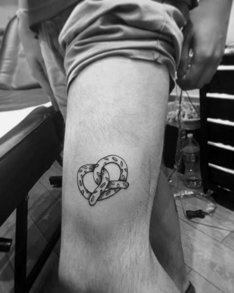 Small black and grey pretzel tattoo on the upper thigh, featuring fine linework and subtle shading for a minimalist yet detailed look.