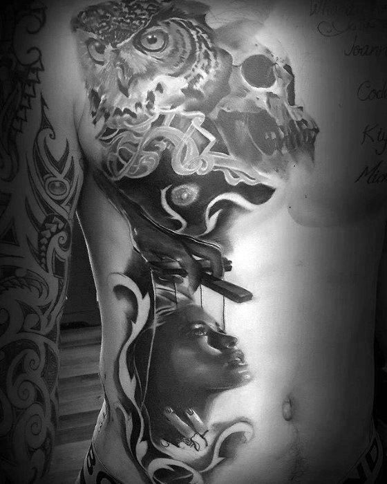 Detailed black and grey puppet tattoo featuring an owl, skull, and a hand controlling a woman’s face with strings, symbolizing wisdom and control.