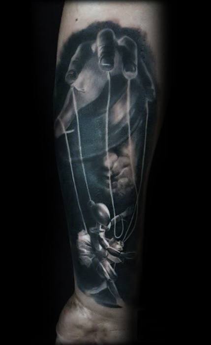Realistic black and grey puppet tattoo of a large hand controlling a ballerina marionette, symbolizing control, fragility, and grace.