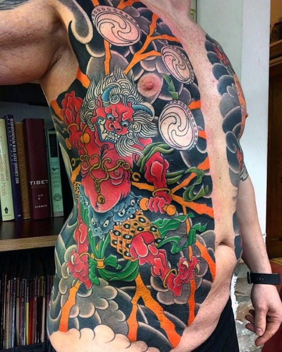 Vibrant Raijin side torso tattoo with red thunder god, orange lightning, swirling clouds, and traditional drums in bold Japanese style.