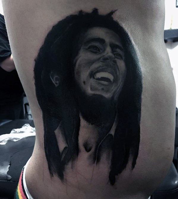 Black and gray rib tattoo of Bob Marley smiling, with detailed shading and long dreadlocks flowing down for a bold portrait look.