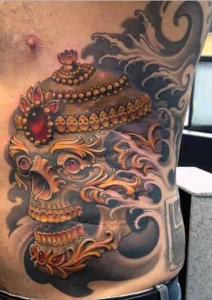 Side torso Tibetan skull tattoo with golden crown, red jewel, swirling smoke, and rich orange and gold ornamental details.