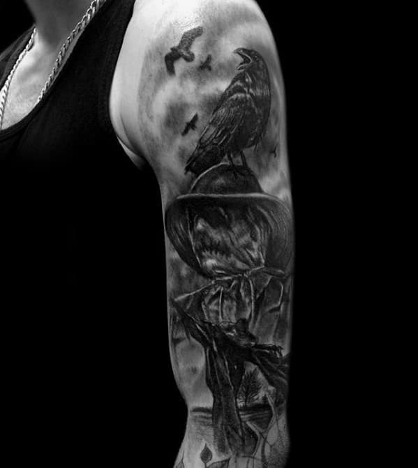 Realistic black and grey scarecrow tattoo with a perched crow, flying birds, full moon, and eerie shadowed details on the upper arm.