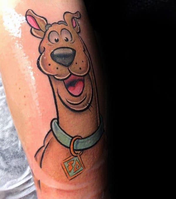 Tattoo of Scooby-Doo smiling brightly with wide eyes, wearing his signature teal collar and gold tag on a forearm.