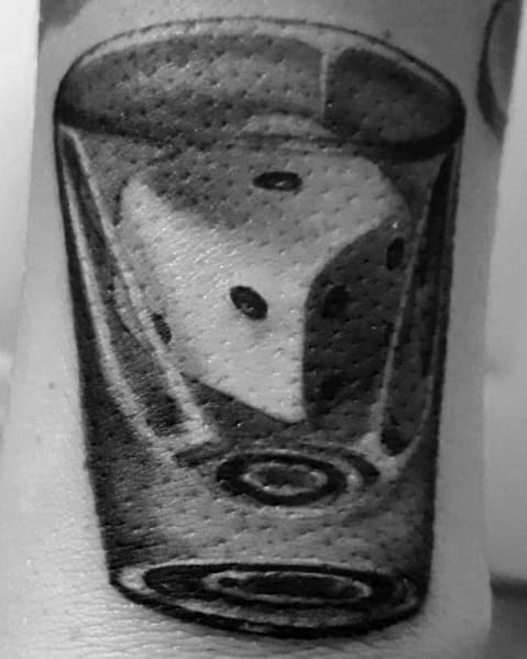 Black and grey shot glass tattoo with a detailed die inside, creating a bold and realistic gambling-inspired design with smooth shading.
