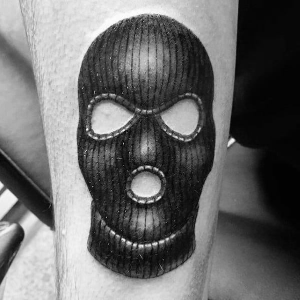 Tattoo of a black ski mask with bold vertical lines and wide eye and mouth openings, placed on the lower leg.