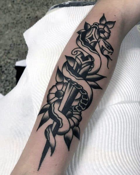 Black and grey snake and dagger tattoo on forearm, featuring bold shading, floral elements, and a striking traditional design.