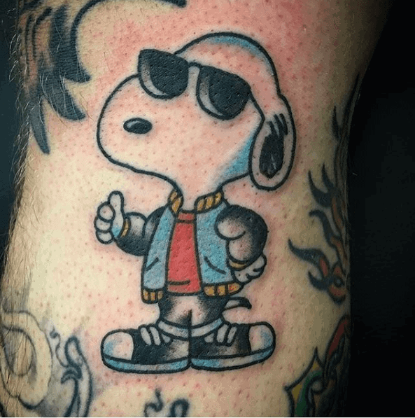 Colorful tattoo of Snoopy wearing sunglasses, a blue jacket, red shirt, and sneakers, giving a thumbs-up, placed on a person's leg.