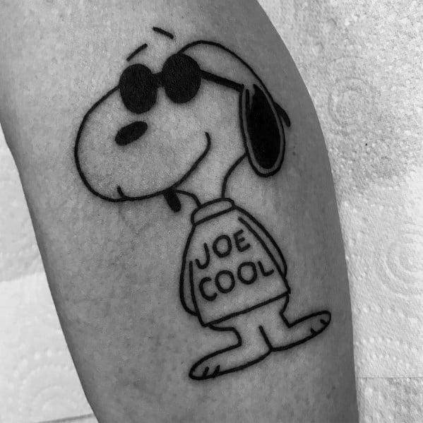 Black and white tattoo of Snoopy as "Joe Cool," wearing sunglasses and a sweater with "Joe Cool" written on it, placed on a person's arm.