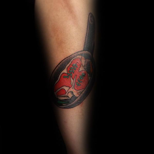 Tattoo of a sizzling steak in a frying pan, with vibrant red tones and seasoning details, creating a bold and dynamic cooking-inspired design